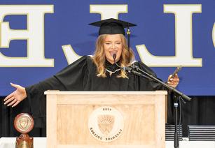 Student Rosie Luke wearing graduation regalia while standing at podium spreading her arms wide in celebration